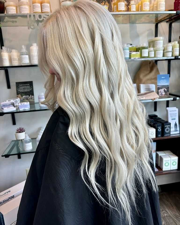 Woman with long, wavy, platinum blonde hair in a salon, surrounded by hair care products.