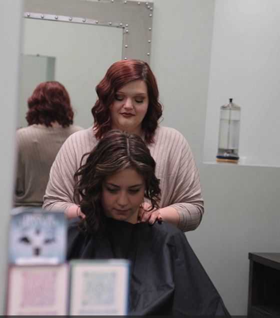 A stylist with red hair standing behind a seated client in a salon, adjusting her hair.